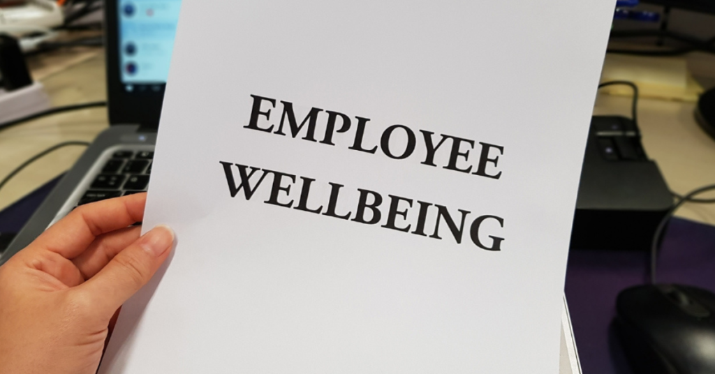 Obrazek przedstawia kartke z napisem "Employee Wellbeing", trzymaną przez osobę. w tle komputer i przestrzeń biurowa.
